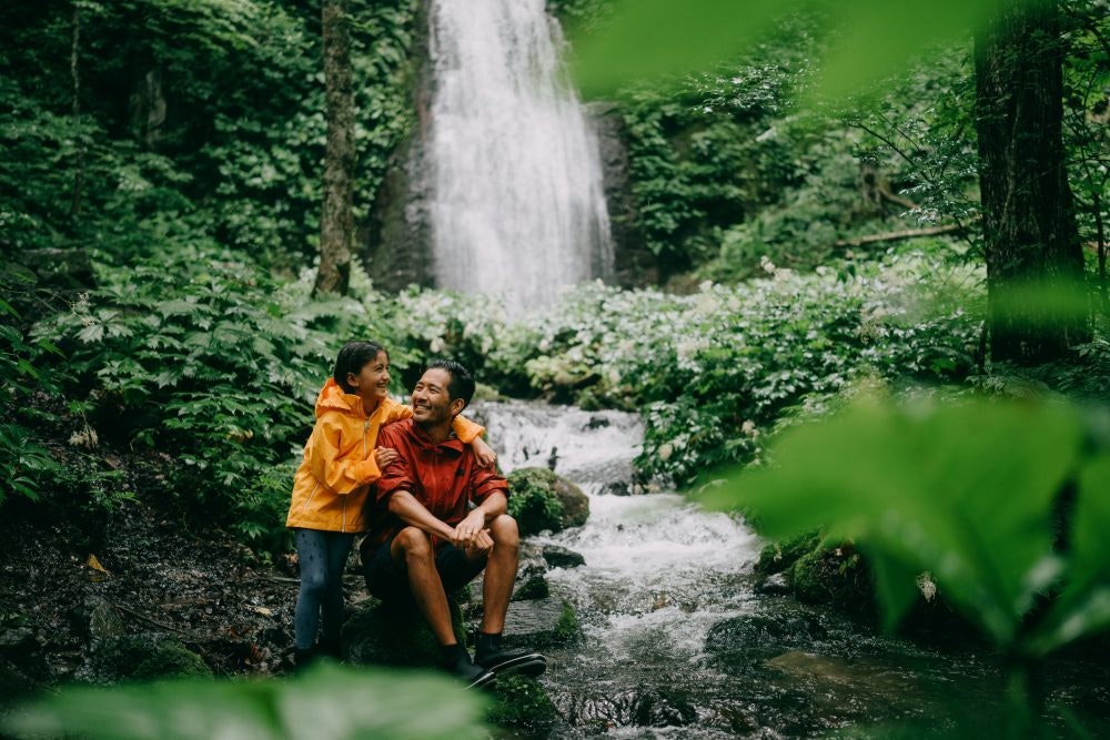 A father and child sit smiling in nature next to a waterfall